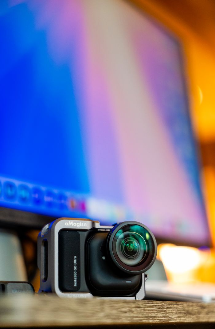 about-us-01 Close-up of a compact digital camera on a desk with a blurred computer screen in the background.