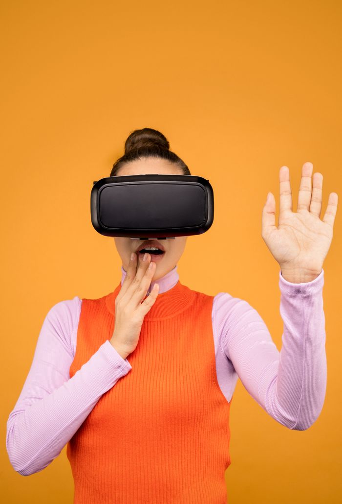 about-us-02 Woman enjoying a virtual reality experience wearing a VR headset against a vibrant orange background.
